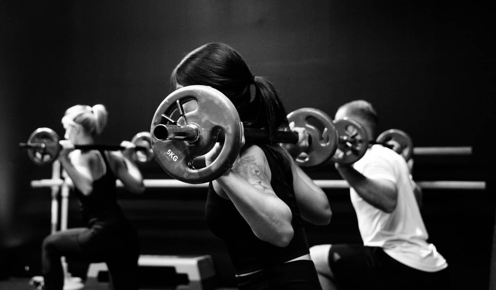 Three training partners squatting barbells side by side in a dark gym, black and white